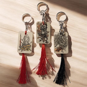 Puede incluir: Tres llaveros de madera con diferentes ilustraciones y borlas. El primer llavero presenta un personaje con una corona y una espada, el segundo llavero presenta un personaje con una corona y una espada, y el tercer llavero presenta un personaje con una corona y una espada. Las borlas son rojas y negras.