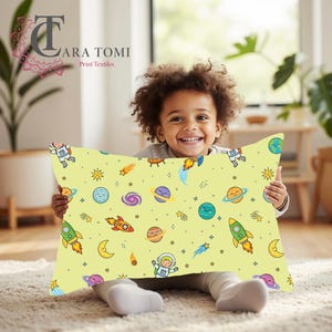 May include: A rectangular pillow with a light yellow background featuring a repeating pattern of cartoon astronauts, colourful planets with faces, rockets, stars, and crescent moons. The design includes various celestial bodies in shades of blue, orange, purple, and green.