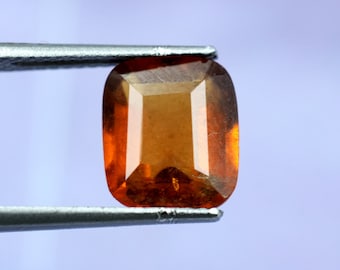 May include: A close-up of a brown gemstone with a rectangular cut. The gemstone is held in place by a pair of tweezers.