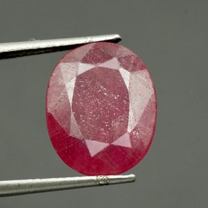 May include: Oval-cut ruby gemstone, held by silver tweezers. The gem has a deep red color with multiple facets that reflect light. The ruby is a precious stone, often used in jewelry.