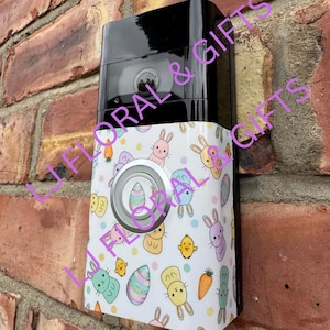 May include: A white doorbell cover with a cute bunny and Easter egg design. The cover has a light pink, light blue, and yellow color scheme. The cover is designed to fit a Ring doorbell.