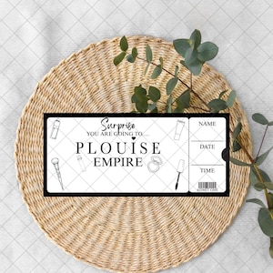 May include: A black and white ticket design with the text "Surprise You are going to... PLOUISE EMPIRE". The ticket includes spaces for name, date, and time, along with illustrations of makeup items. The ticket is placed on a woven mat.