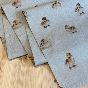 May include: A linen table runner featuring a repeated pattern of brown ducklings. The runner is folded in thirds, showcasing the design on all three layers.
