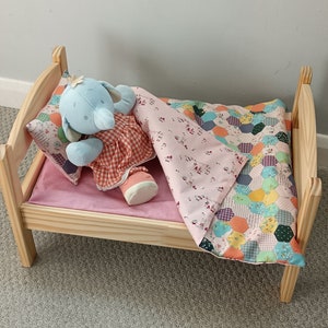May include: A small wooden doll bed with a light pink mattress, a patterned pillow, and a patchwork quilt. A blue elephant doll wearing a red and white checkered dress is lying on the bed.