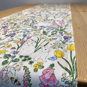 May include: White table runner with a floral pattern featuring bunnies, butterflies, and flowers in shades of pink, yellow, blue, and purple.