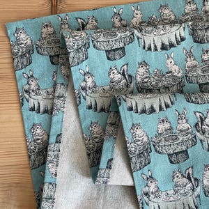 May include: A blue and white fabric with a repeating pattern of squirrels, rabbits, and teacups. The fabric has a subtle texture and a vintage feel.
