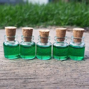 Könnte beinhalten: Fünf kleine Glasflaschen mit Korkstopfen, gefüllt mit einer leuchtend grünen Flüssigkeit. Die Flaschen sind in einer Reihe auf einer Holzoberfläche angeordnet.