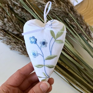 Puede incluir: Adorno blanco en forma de corazón con bordado floral azul y hojas verdes. Se adjunta una cinta blanca para colgarlo. El adorno mide aproximadamente 10 cm de largo. Artículo decorativo para la decoración del hogar.