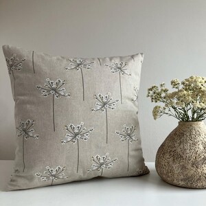 Op de afbeelding: Een vierkant beige sierkussen met een herhaald patroon van gestileerde witte en grijze bloemillustraties. Het kussen ligt op een wit oppervlak naast een getextureerde vaas met gedroogde bloemen.