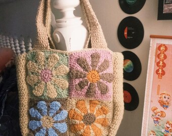 May include: A crocheted tote bag with a floral design. The bag is made of beige yarn and features four different flower motifs in pink, blue, green, and orange.