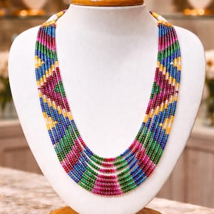 Puede incluir: Collar de múltiples hilos exhibido en un maniquí blanco. El collar presenta una vibrante gama de colores, incluyendo cuentas rojas, verdes, azules y amarillas. Las cuentas están dispuestas en un diseño estampado, creando un accesorio visualmente llamativo.