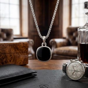 May include: A silver necklace with a square, black onyx pendant hangs in front of a blurred background. A black leather wallet, a vintage wristwatch, and a decanter with dark liquid are on a wooden table.