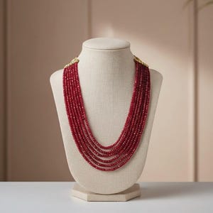 Könnte beinhalten: Mehrreihige Halskette auf einer Schaufensterpuppe ausgestellt. Die Halskette besteht aus zahlreichen Strängen kleiner, roter Perlen. Die Perlen fallen kaskadenartig herab und erzeugen einen vollen und eleganten Look. Die Halskette wird auf einer beigefarbenen Schaufensterpuppe präsentiert.