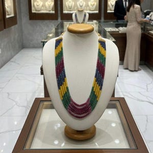 May include: A colourful multi-strand necklace on a display bust. The necklace has strands of yellow, green, red, and blue beads. The jewellery is displayed in a shop, with other necklaces visible in the background.