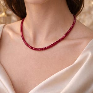 Puede incluir: Primer plano de un collar de cuentas rojas sobre el cuello de una persona. El collar está compuesto por pequeñas cuentas redondas. La persona lleva una blusa de satén color crema. El fondo es un oro suave y borroso.