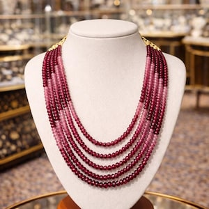 Puede incluir: Collar de múltiples hilos exhibido en un maniquí. El collar presenta hilos de pequeñas cuentas redondas en varios tonos de rojo y rosa. El collar se exhibe en un maniquí blanco con una base de madera, sobre un fondo borroso.