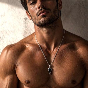May include: A silver necklace with a black pendant hangs against a tanned chest. The pendant is a black triangle shape with a silver border. The person has dark hair and a beard.