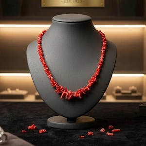 May include: A striking red coral necklace presented on a black jewellery bust. The necklace is made of irregular, cylindrical coral pieces. The background features the brand name "CORALLIA" and the establishment year "EST. 1923".