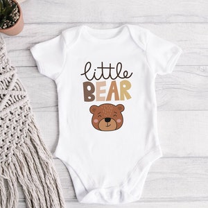 May include: White baby onesie with the text "little bear" in brown and beige lettering. A cartoon brown bear face is below the text.