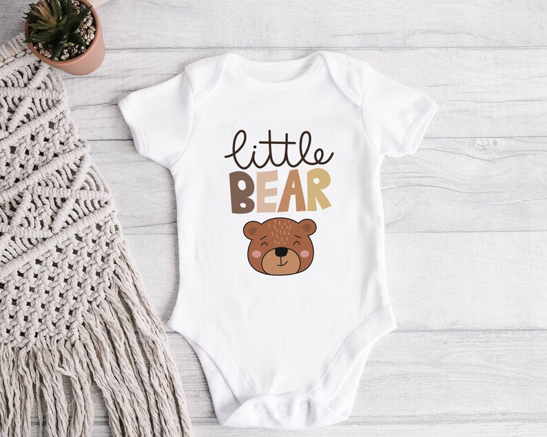 May include: White baby onesie with the text "little bear" in brown and beige lettering. A cartoon brown bear face is below the text.