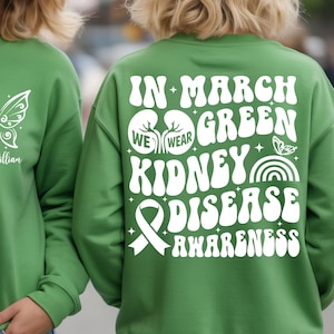 Puede incluir: Sudadera verde con texto y gráficos blancos. La parte delantera presenta un diseño de mariposa y las palabras "Team William". La parte trasera muestra "En marzo usamos la concienciación sobre la enfermedad renal verde" con gráficos de riñones y cintas.