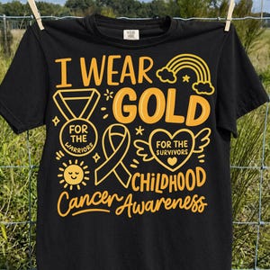 May include: A black short-sleeve t-shirt featuring yellow graphic text and illustrations. The design includes the words I WEAR GOLD, a medal, a ribbon, a heart with wings, a sun, and a rainbow, with the text CHILDHOOD Cancer Awareness.