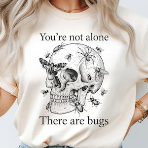 以下が含まれることがあります： 様々な昆虫に囲まれた頭蓋骨の、詳細な白黒イラストが特徴のクリーム色のTシャツ。「You're not alone」の文字が頭蓋骨の上に、「There are bugs」が下に書かれています。ゴシックをテーマにしたグラフィックTシャツ。
