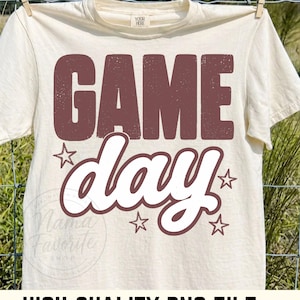 Op de afbeelding: Een crèmekleurig T-shirt met korte mouwen met de tekst GAME in grote, verweerde bruine blokletters boven het woord day in wit cursief schrift. Vier kleine bruine sterren zijn rond de tekst op de voorkant van het kledingstuk geplaatst.
