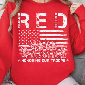 May include: Red sweatshirt with a white graphic of the American flag and soldiers. The text on the sweatshirt reads "REMEMBER EVERYONE DEPLOYED" and "HONORING OUR TROOPS".