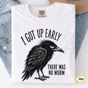 May include: A white short-sleeve t-shirt featuring a black graphic illustration of a grumpy crow. The text above the crow reads I GOT UP EARLY and the text below the crow reads THERE WAS NO WORM. The shirt has a Comfort Colors brand label at the neck.