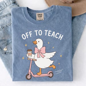 May include: A blue crew neck t-shirt featuring a white goose wearing a pink bow, holding a coffee cup, and riding a pink scooter. The text "OFF TO TEACH" is printed in white above the goose, surrounded by small yellow stars on a blue fabric.