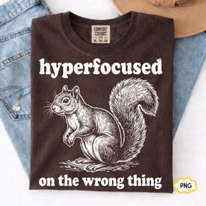 Puede incluir: Una camiseta de color marrón oscuro con una ilustración de líneas blancas de una ardilla en el centro. El texto "hyperfocused" está impreso sobre la ardilla y el texto "on the wrong thing" debajo en una fuente serif blanca y negrita.