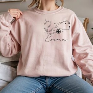 May include: A light pink sweatshirt featuring a black stethoscope design and the name "Emma" in cursive script. The design includes small star accents. The sweatshirt is a solid color and has long sleeves.