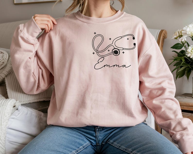 May include: A light pink sweatshirt featuring a black stethoscope design and the name "Emma" in cursive script. The design includes small star accents. The sweatshirt is a solid color and has long sleeves.