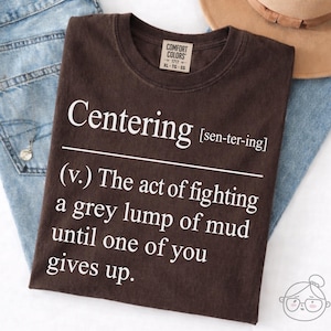 May include: A brown t-shirt with the word "Centering" in white, followed by a definition. The definition reads: "(v.) The act of fighting a grey lump of mud until one of you gives up."
