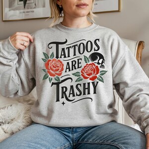 May include: A light gray sweatshirt with the text "Tattoos are Trashy" in black script, adorned with red roses and a black skull. The design is centered on the chest of the sweatshirt.