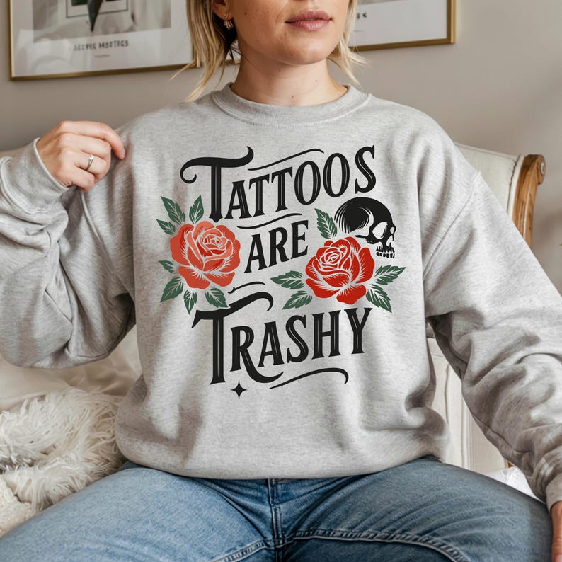 May include: A light gray sweatshirt with the text "Tattoos are Trashy" in black script, adorned with red roses and a black skull. The design is centered on the chest of the sweatshirt.