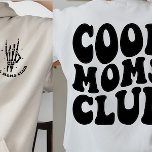 May include: Two white hoodies with black graphic designs. One hoodie features a skeleton hand with the text "Cool Moms Club". The other hoodie features the text "Cool Moms Club" in a retro font.