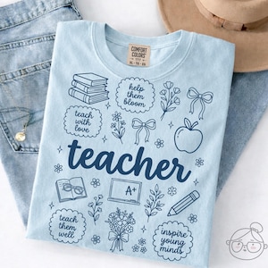 Könnte beinhalten: Hellblaues Comfort Colors T-Shirt mit dem Wort "teacher" in dunkelblauer Schrift. Das Shirt zeigt verschiedene Illustrationen zum Thema Schule, darunter Bücher, einen Apfel und einen Bleistift. Größe XL-TG-EG.