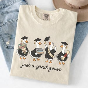 May include: Cream-colored t-shirt featuring a whimsical illustration of four geese in graduation attire. The geese are depicted with graduation caps, gowns, and holding items like flowers, books, and a diploma. The text "just a grad goose" is written in a script font.