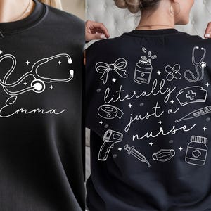 May include: Black sweatshirt with white graphic designs. The front features a stethoscope and the name "Emma". The back displays medical-themed illustrations and the phrase "literally just a nurse".