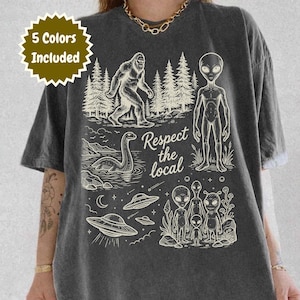 May include: Dark gray t-shirt with a graphic design featuring Bigfoot, the Loch Ness Monster, aliens, and UFOs. The text "Respect the local" is also included in the design. The shirt has a vintage, worn look.