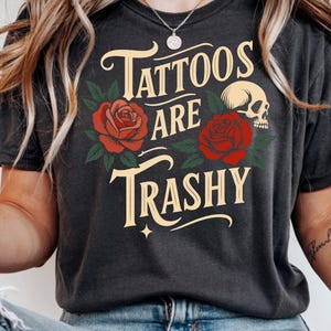 May include: Dark gray t-shirt with the text "Tattoos are Trashy" in a vintage font, accompanied by red roses, green leaves, and a skull graphic. The shirt has a crew neck and short sleeves.