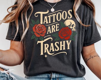 May include: Dark gray t-shirt with the text "Tattoos are Trashy" in a vintage font, accompanied by red roses, green leaves, and a skull graphic. The shirt has a crew neck and short sleeves.