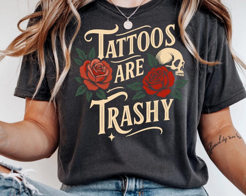 May include: Dark gray t-shirt with the text "Tattoos are Trashy" in a vintage font, accompanied by red roses, green leaves, and a skull graphic. The shirt has a crew neck and short sleeves.