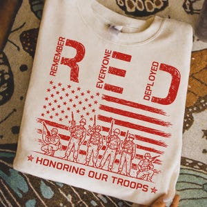 K&ouml;nnte beinhalten: Cremefarbenes Sweatshirt mit einem roten Used-Look-Grafik. Das Design zeigt das Wort "RED" mit den Buchstaben "Remember Everyone Deployed", eine amerikanische Flagge und Soldaten. Der Text "HONORING OUR TROOPS" ist ebenfalls enthalten.