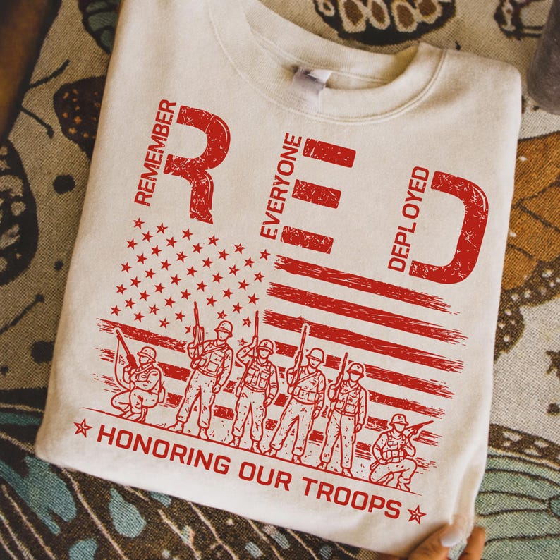 K&ouml;nnte beinhalten: Cremefarbenes Sweatshirt mit einem roten Used-Look-Grafik. Das Design zeigt das Wort "RED" mit den Buchstaben "Remember Everyone Deployed", eine amerikanische Flagge und Soldaten. Der Text "HONORING OUR TROOPS" ist ebenfalls enthalten.