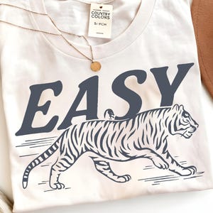 May include: Cream-colored t-shirt with the word "EASY" in dark blue block letters above a tiger illustration. The tiger is also dark blue with white stripes. A gold necklace and a brown hat are visible.