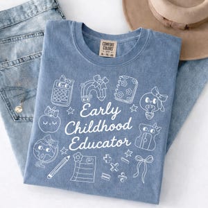 May include: A blue Comfort Colors t-shirt with the words "Early Childhood Educator" and various school-themed graphics in white. The shirt is folded on top of a pair of blue jeans.