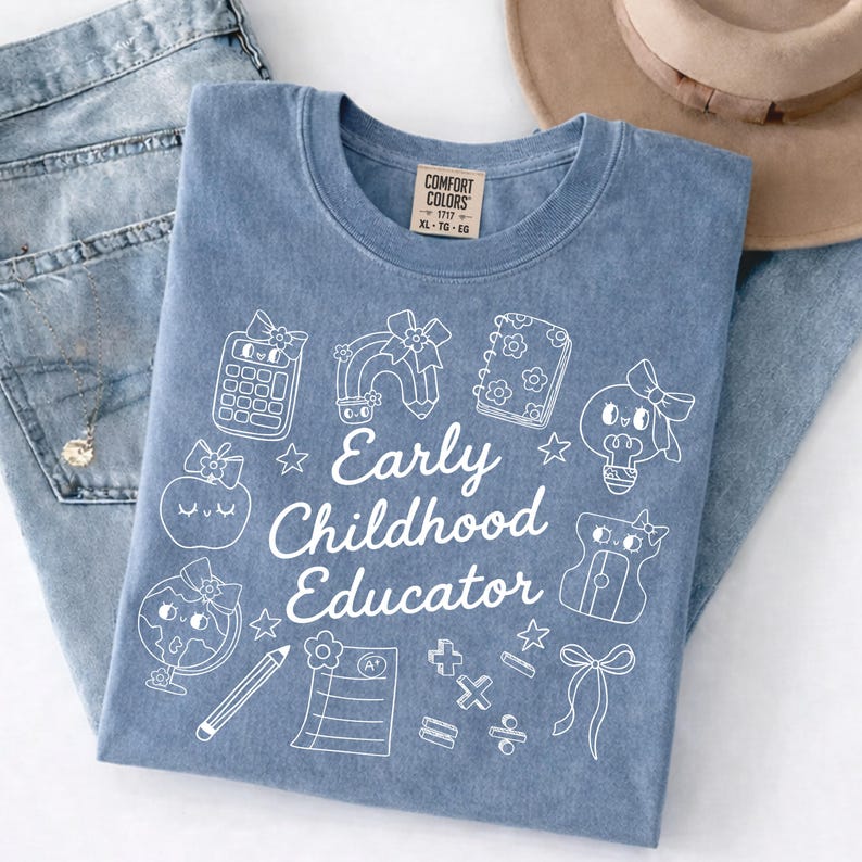 May include: A blue Comfort Colors t-shirt with the words "Early Childhood Educator" and various school-themed graphics in white. The shirt is folded on top of a pair of blue jeans.